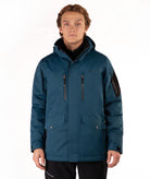 A man wearing a dark teal hooded outdoor jacket with black zippers, flap pockets, and a black sleeve patch, photographed against a plain white studio background.