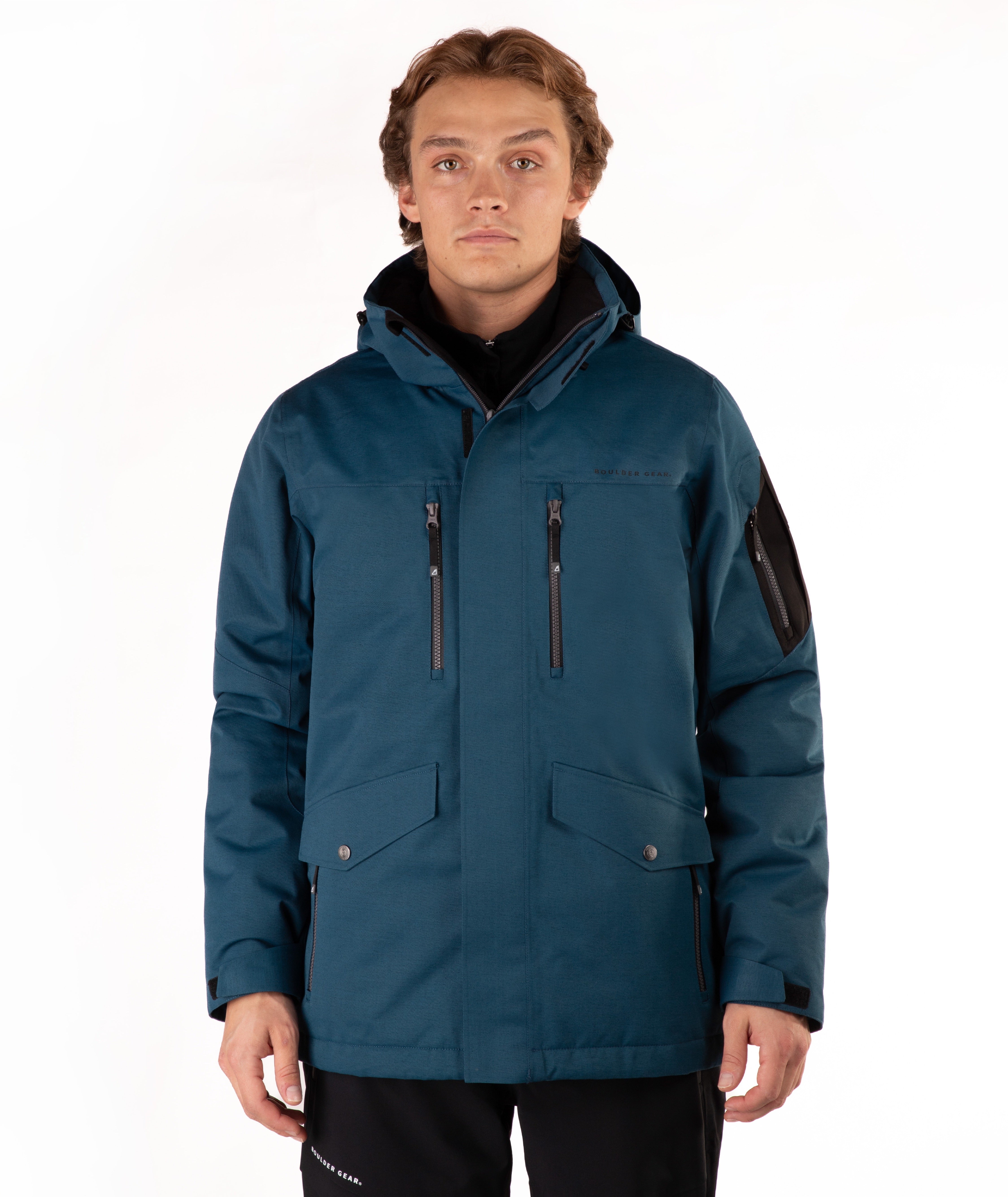 A man wearing a dark teal hooded outdoor jacket with black zippers, flap pockets, and a black sleeve patch, photographed against a plain white studio background.