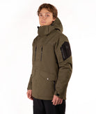 A man wearing an olive green hooded technical jacket with black accents, including a black sleeve patch and black pants, photographed against a clean white studio background.