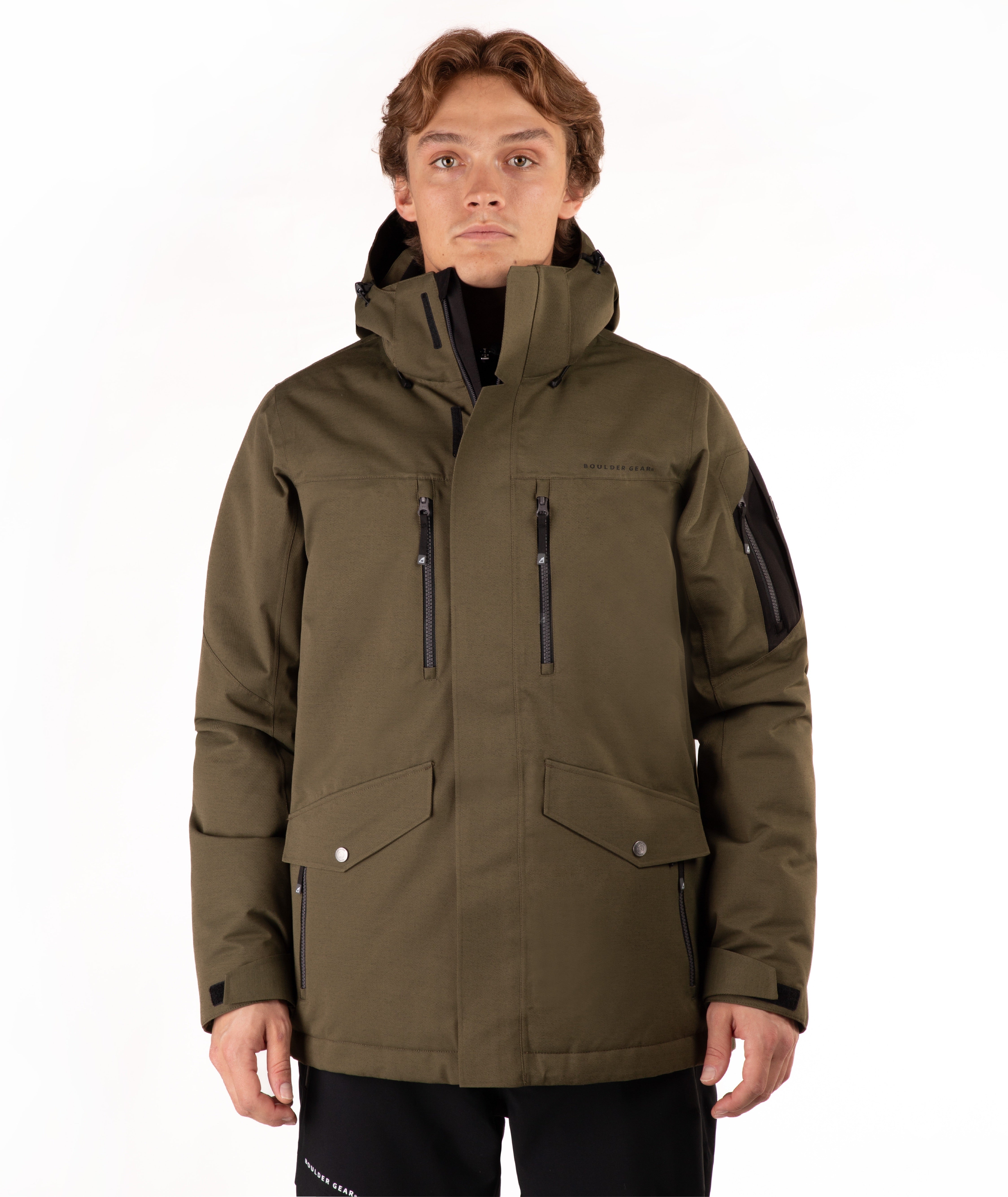 A man wearing an olive green hooded outdoor jacket with black zippers and black pants, photographed against a plain white studio background.