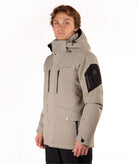 A man wearing a taupe hooded technical jacket with black zippers, black sleeve panels, and a black collar, standing against a plain white background.