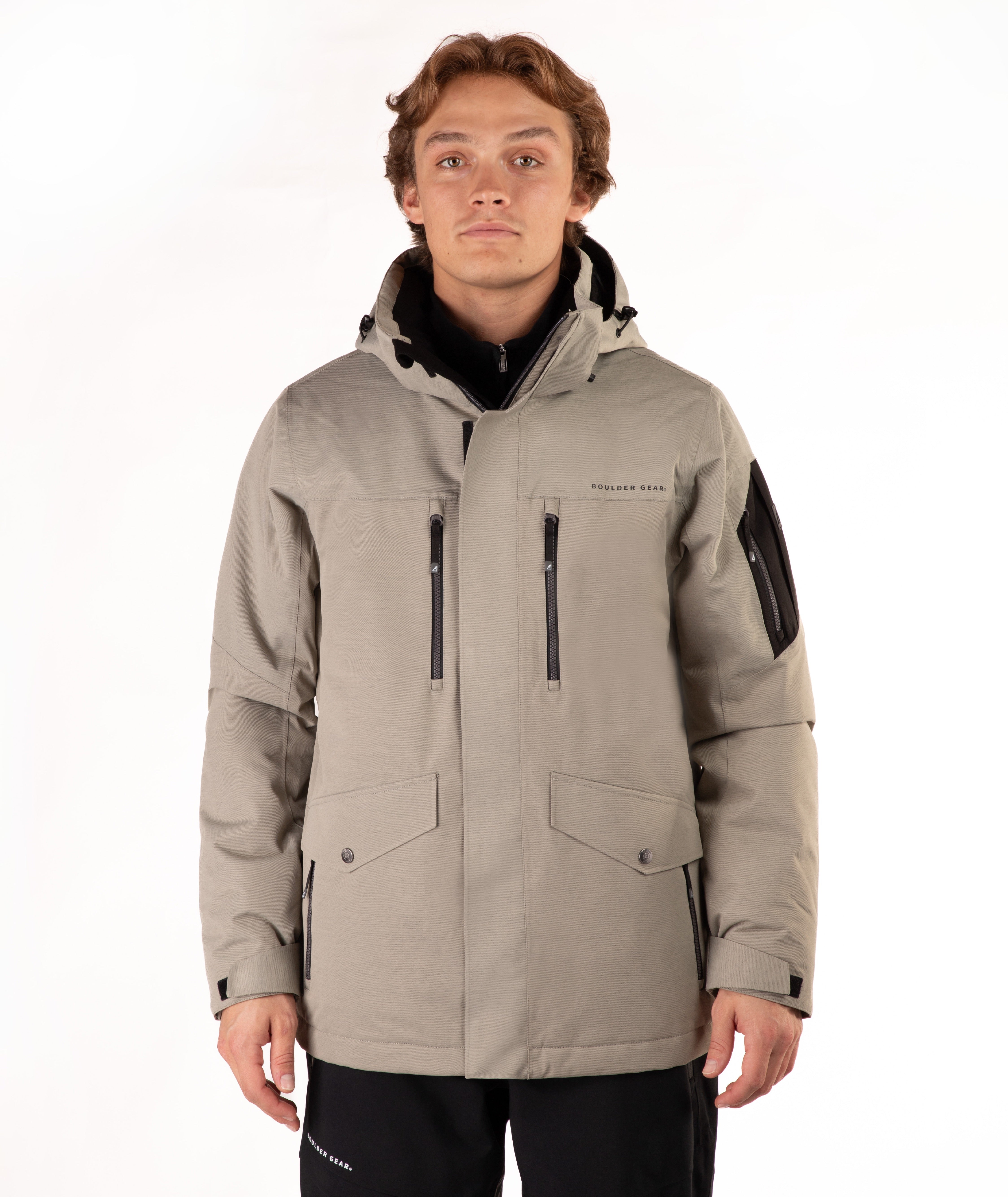 A man wearing a taupe hooded technical jacket with black zippers, black sleeve panels, and a black collar, standing against a plain white background.