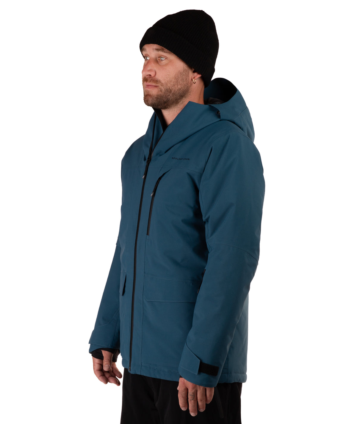 A man wearing a dark teal hooded winter jacket with black zippers, a black beanie, and black pants, photographed against a plain white studio background.
