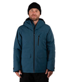 A man wearing a dark teal hooded winter jacket with black zippers, a black beanie, and black pants, photographed against a plain white studio background.