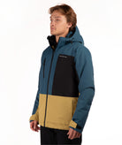 A man wearing a color-blocked outdoor jacket, featuring teal blue, black, and mustard yellow sections, photographed against a plain white studio background.
