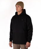 A man wearing a black pullover hoodie with a small square logo patch on the left chest, paired with a brown beanie and khaki pants, photographed against a plain white studio background.