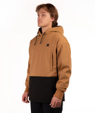 A man wearing a colorblock pullover hoodie with a camel brown upper half and a black lower half, featuring a small black square logo on the left chest, shot against a plain white background.
