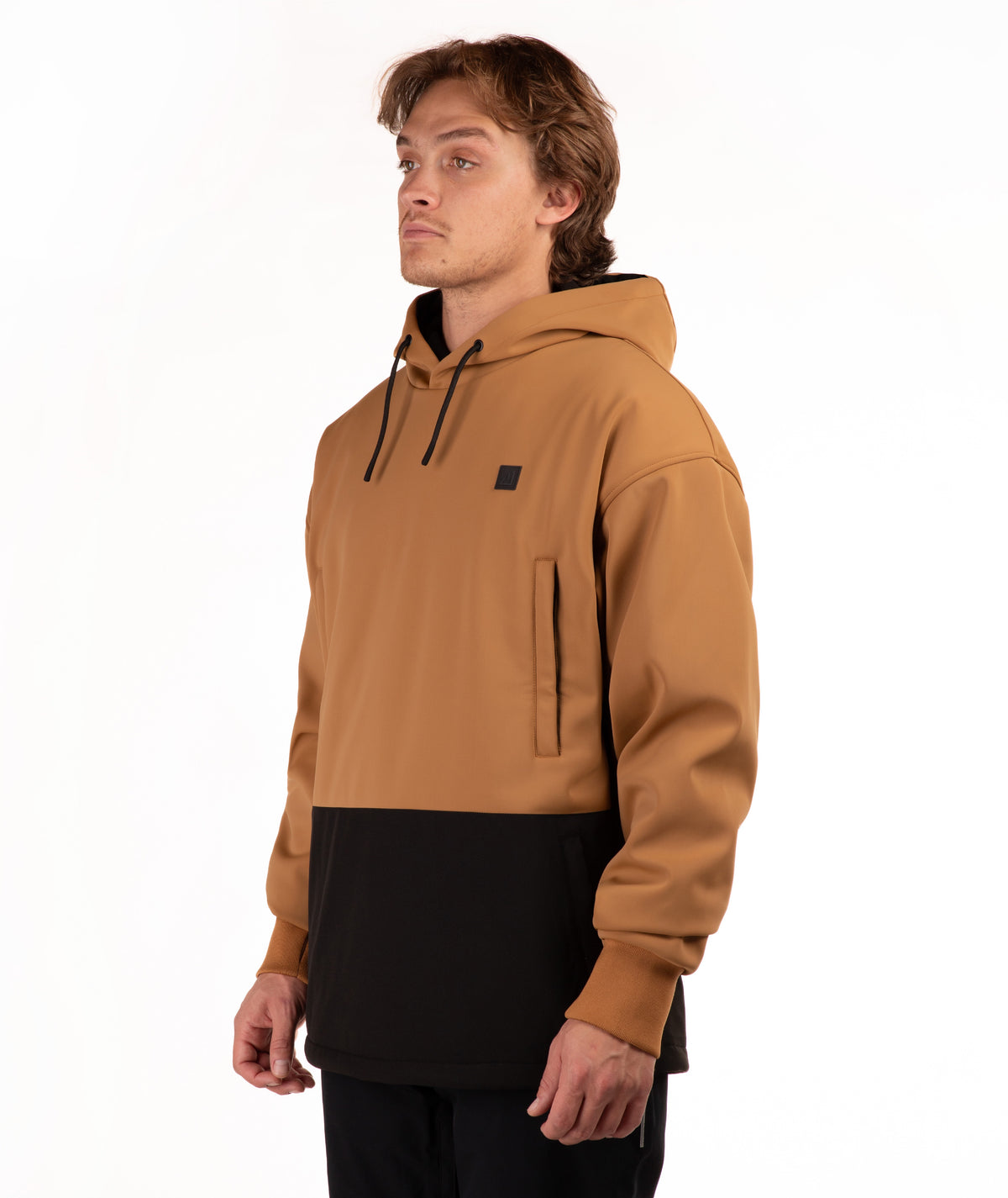 A man wearing a colorblock pullover hoodie with a camel brown upper half and a black lower half, featuring a small black square logo on the left chest, shot against a plain white background.