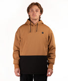 A man wearing a colorblock pullover hoodie with a camel brown upper half and a black lower half, featuring a small black square logo on the left chest, shot against a plain white background.