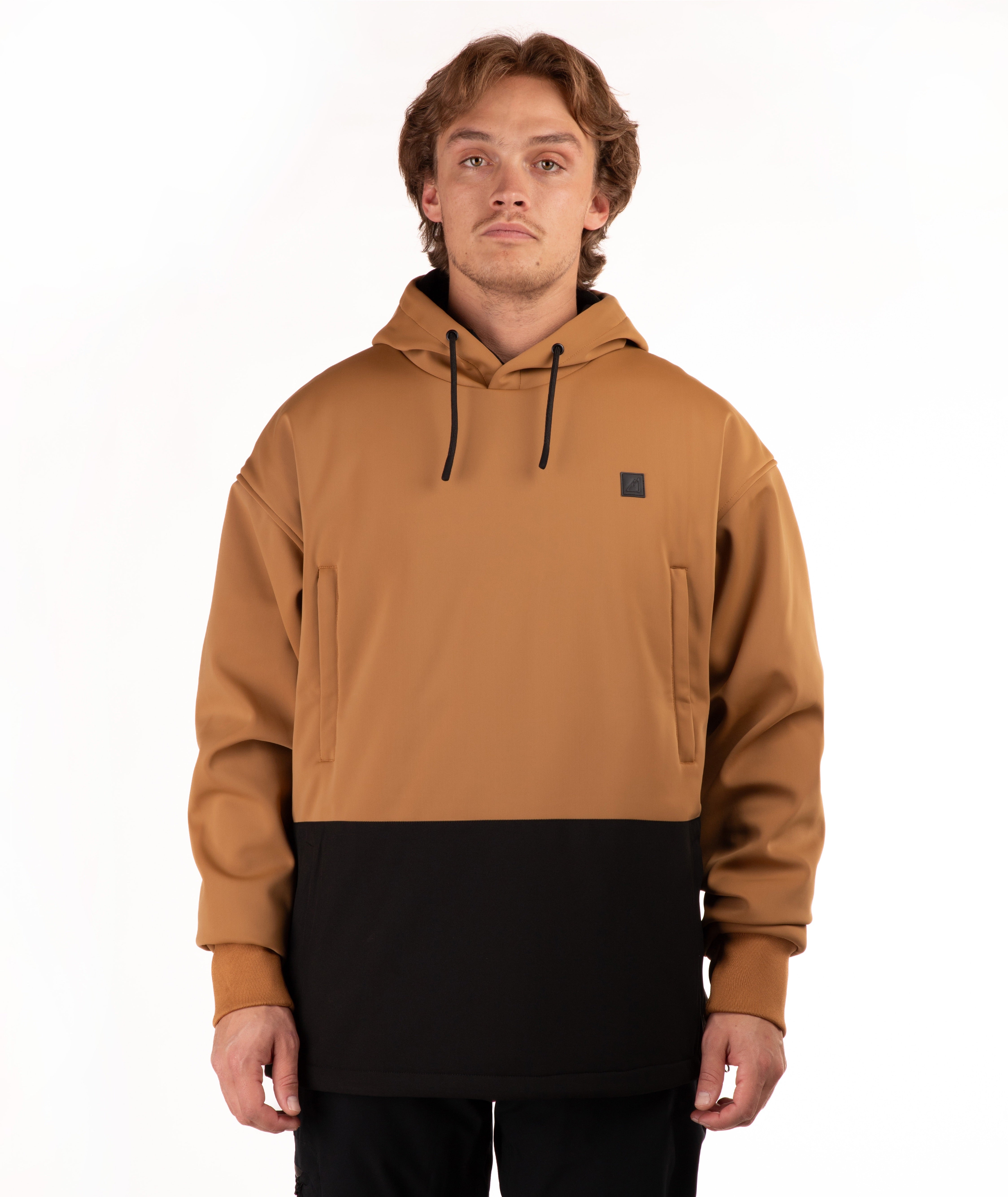 A man wearing a colorblock pullover hoodie with a camel brown upper half and a black lower half, featuring a small black square logo on the left chest, shot against a plain white background.