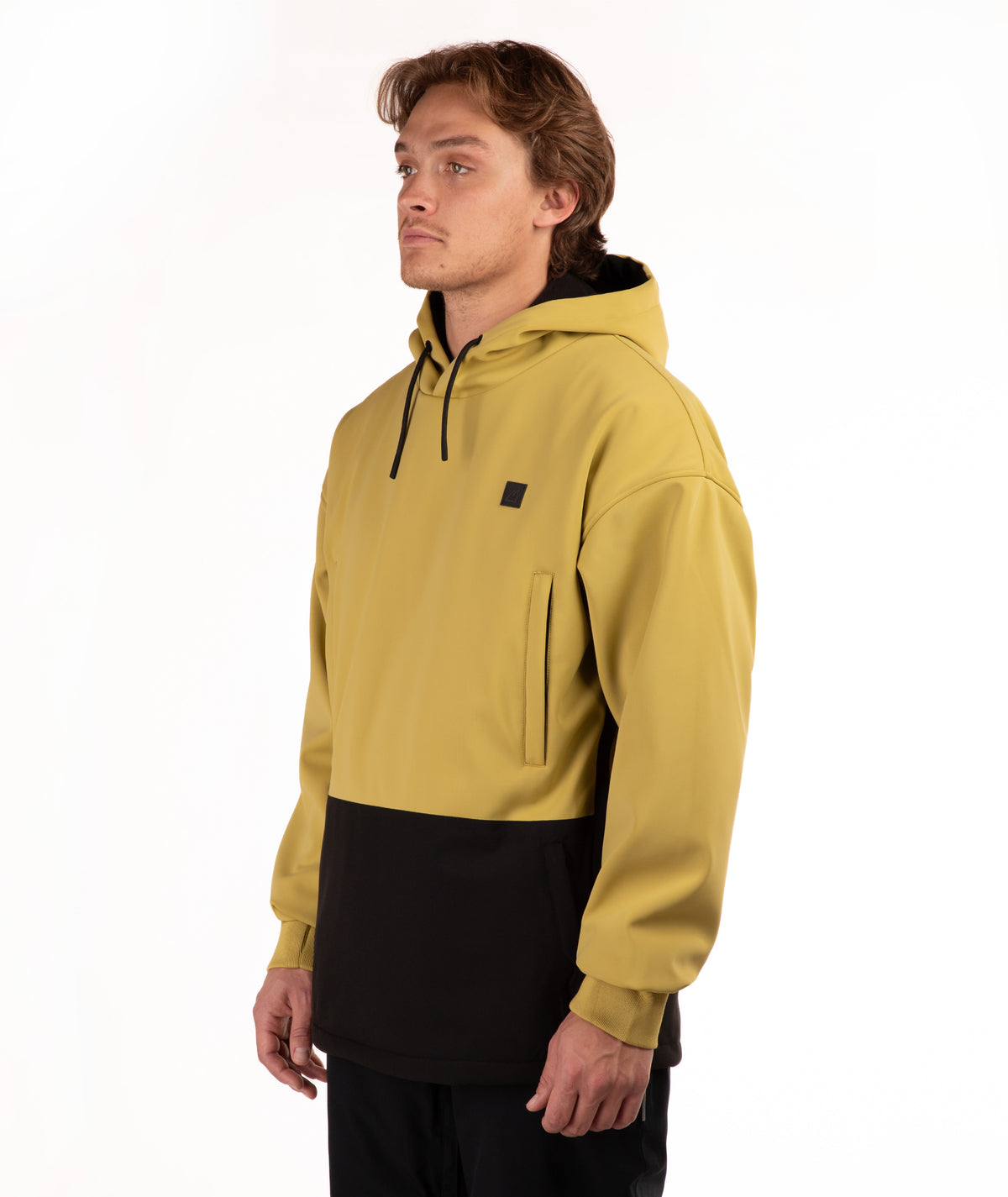 A man wearing a color-block pullover hoodie in mustard yellow and black, featuring a small black square logo on the left chest, photographed against a plain white studio background.