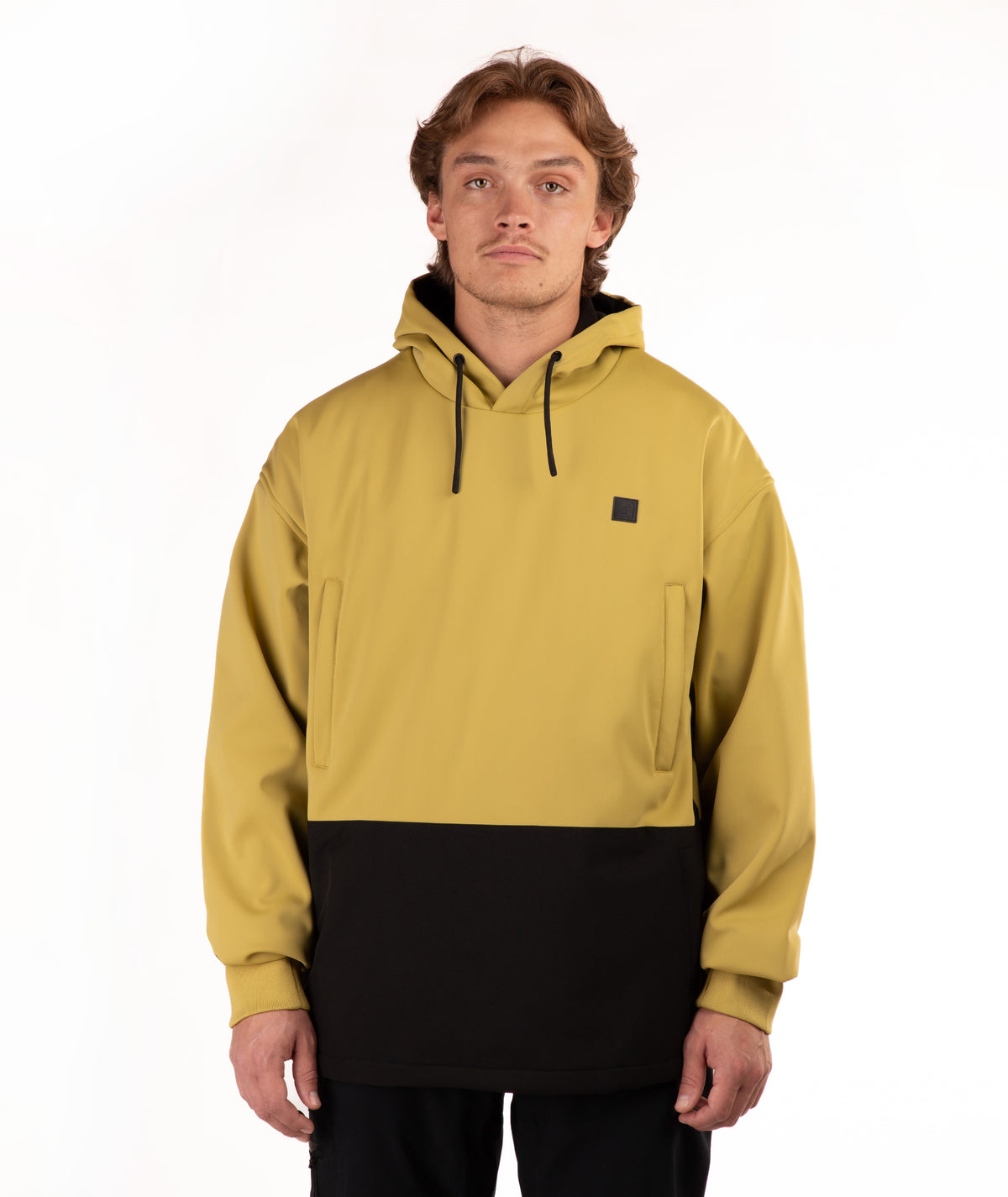 A man wearing a color-block pullover hoodie in mustard yellow and black, featuring a small black square logo on the left chest, photographed against a plain white studio background.