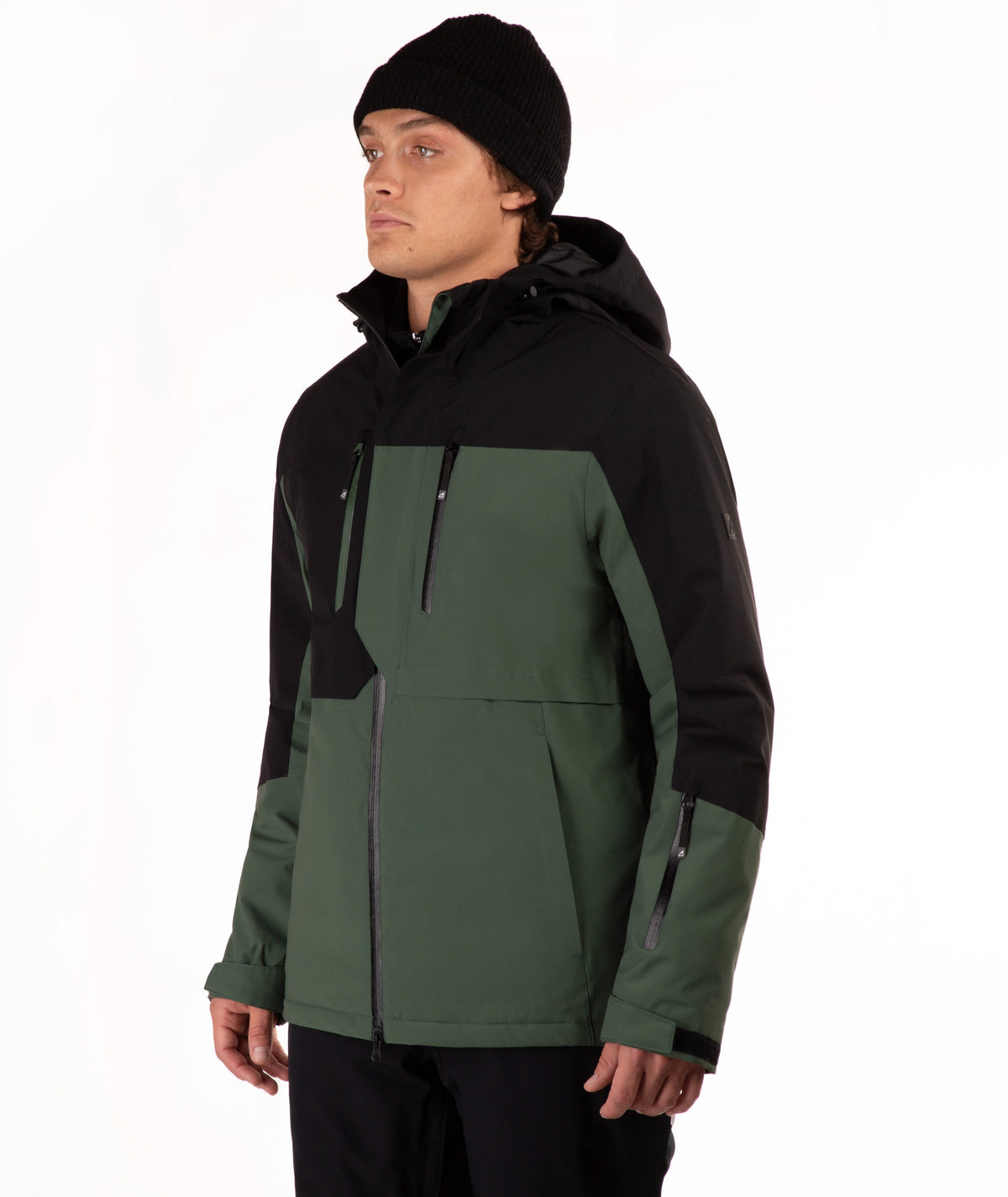 A man wearing a black and dark green color-blocked ski jacket, a black beanie, and black pants, standing against a plain white studio background.