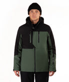 A man wearing a black and dark green color-blocked ski jacket, a black beanie, and black pants, standing against a plain white studio background.