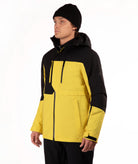 A man wearing a yellow and black ski jacket, black pants, and a black beanie, standing against a plain white studio background.