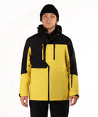 A man wearing a yellow and black ski jacket, black pants, and a black beanie, standing against a plain white studio background.