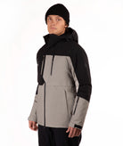 A man wearing a color-blocked ski jacket in black and taupe, paired with a black beanie, photographed against a plain white studio background.