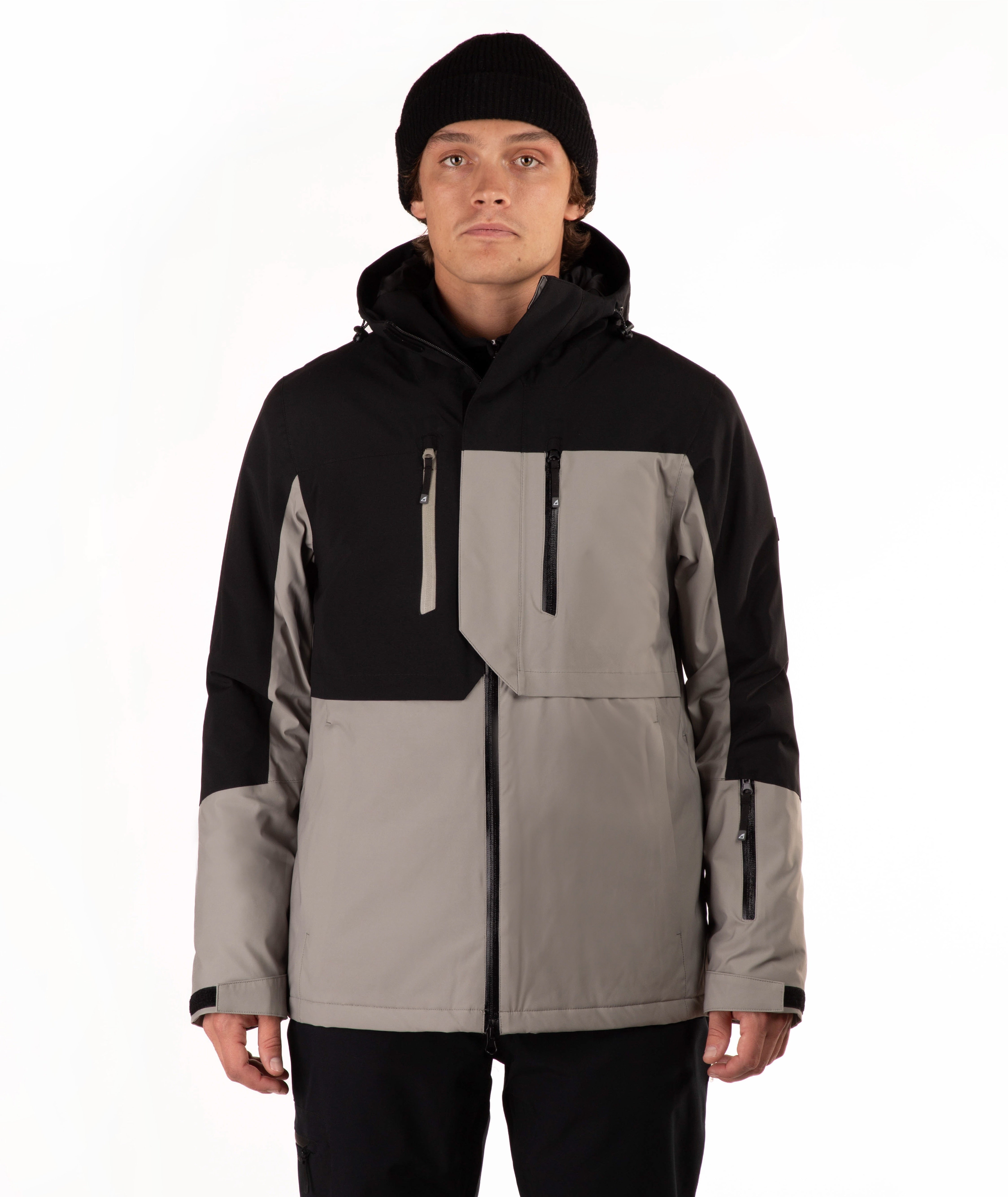 A man wearing a color-blocked ski jacket in black and taupe, paired with a black beanie, photographed against a plain white studio background.