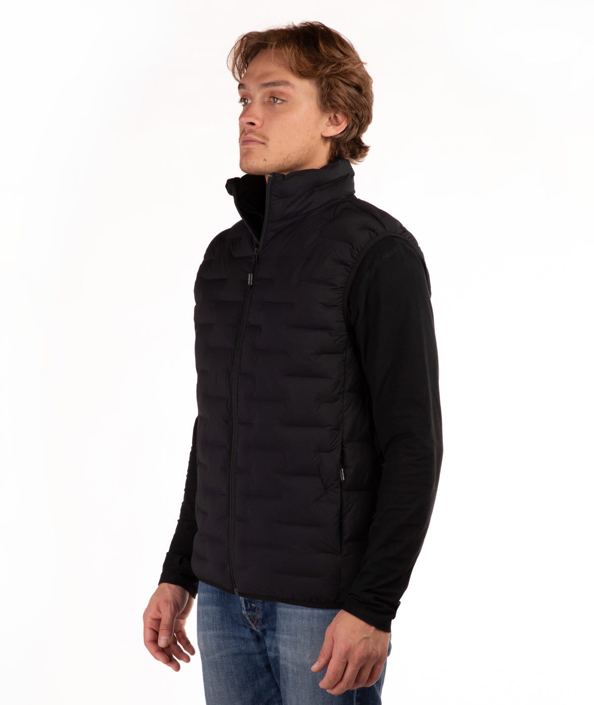 A man wearing a black zip-up puffer vest over a black long-sleeve shirt and blue jeans, standing against a plain white background.