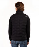 A man viewed from the back wearing a black quilted vest over a black long-sleeve shirt and blue jeans, against a plain white background.