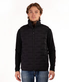 A man wearing a black zip-up puffer vest over a black long-sleeve shirt and blue jeans, standing against a plain white background.