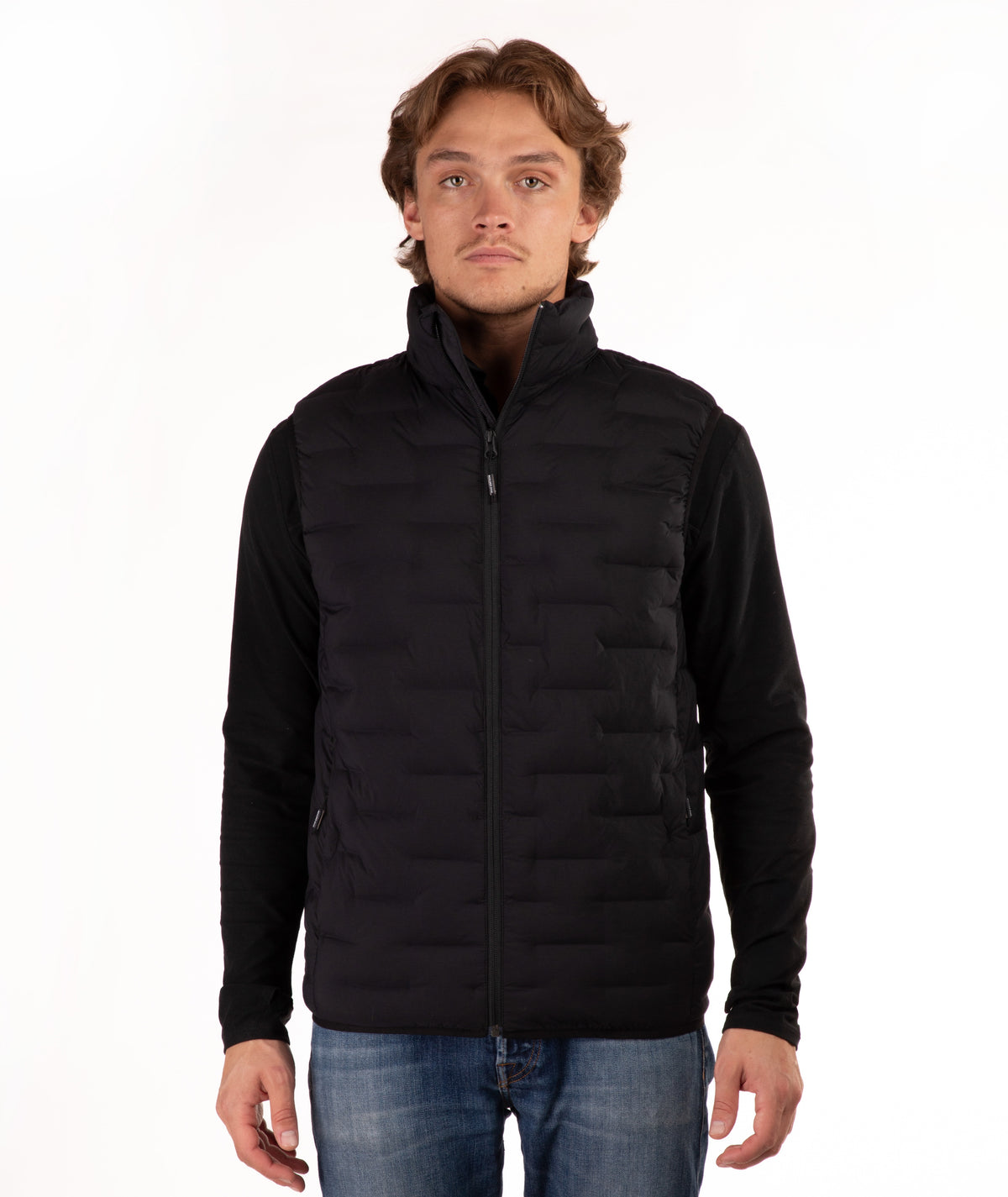 A man wearing a black zip-up puffer vest over a black long-sleeve shirt and blue jeans, standing against a plain white background.