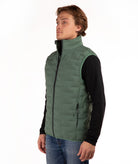 A man wearing a green quilted puffer vest over a black long-sleeve shirt and blue jeans, standing against a plain white background.