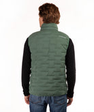 A man viewed from behind wearing a  green quilted puffer vest over a black long-sleeve shirt and blue jeans, against a plain white background.