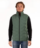 A man wearing a green quilted puffer vest over a black long-sleeve shirt and blue jeans, standing against a plain white background.
