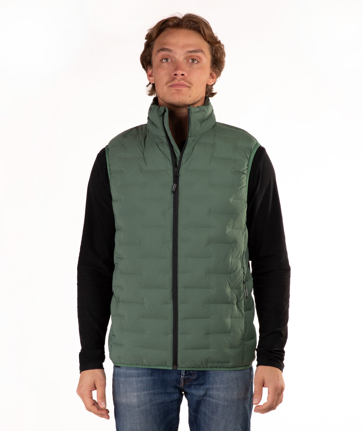 A man wearing a green quilted puffer vest over a black long-sleeve shirt and blue jeans, standing against a plain white background.