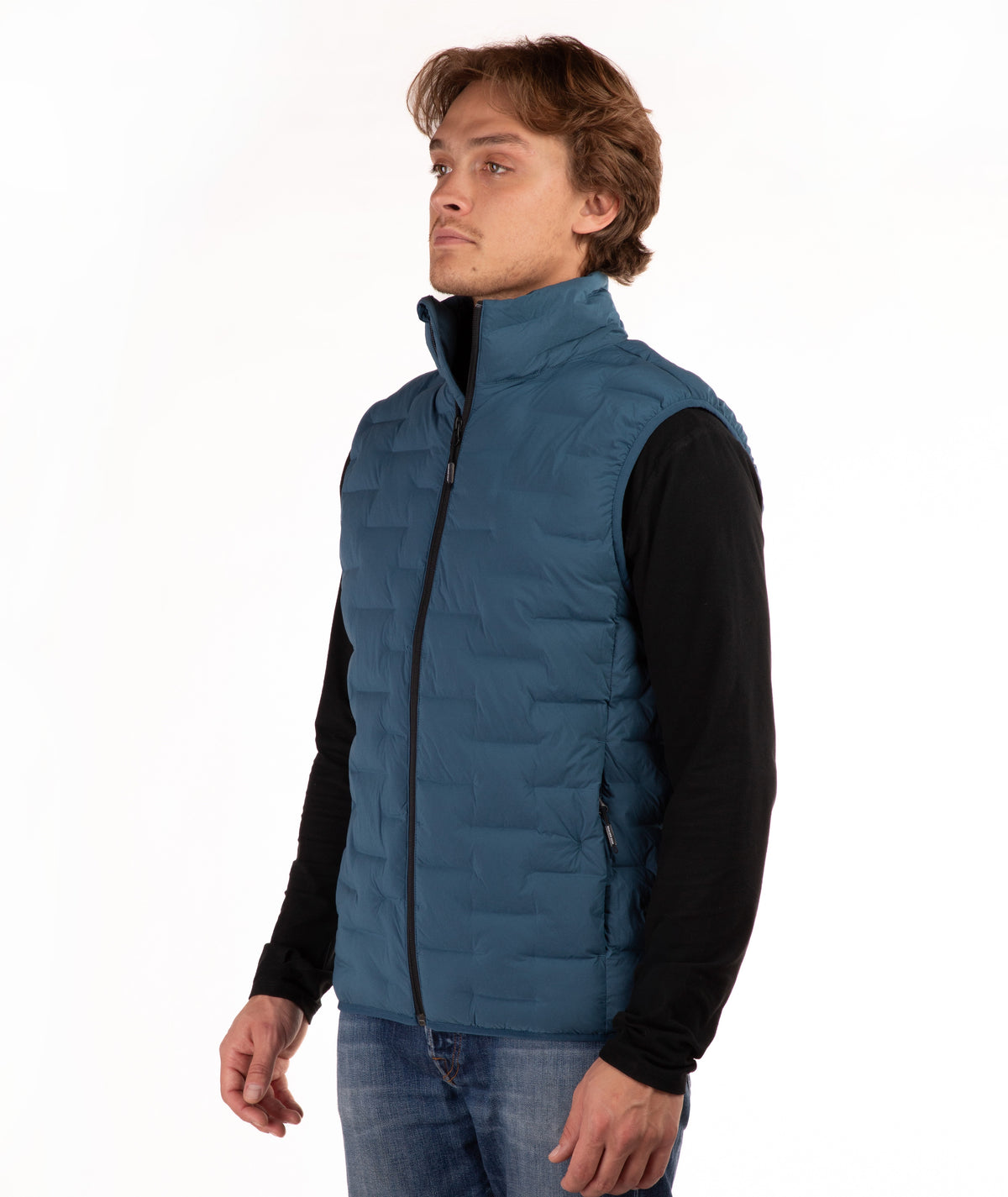 A man wearing a teal blue quilted puffer vest over a black long-sleeve shirt and blue jeans, standing against a plain white studio background.