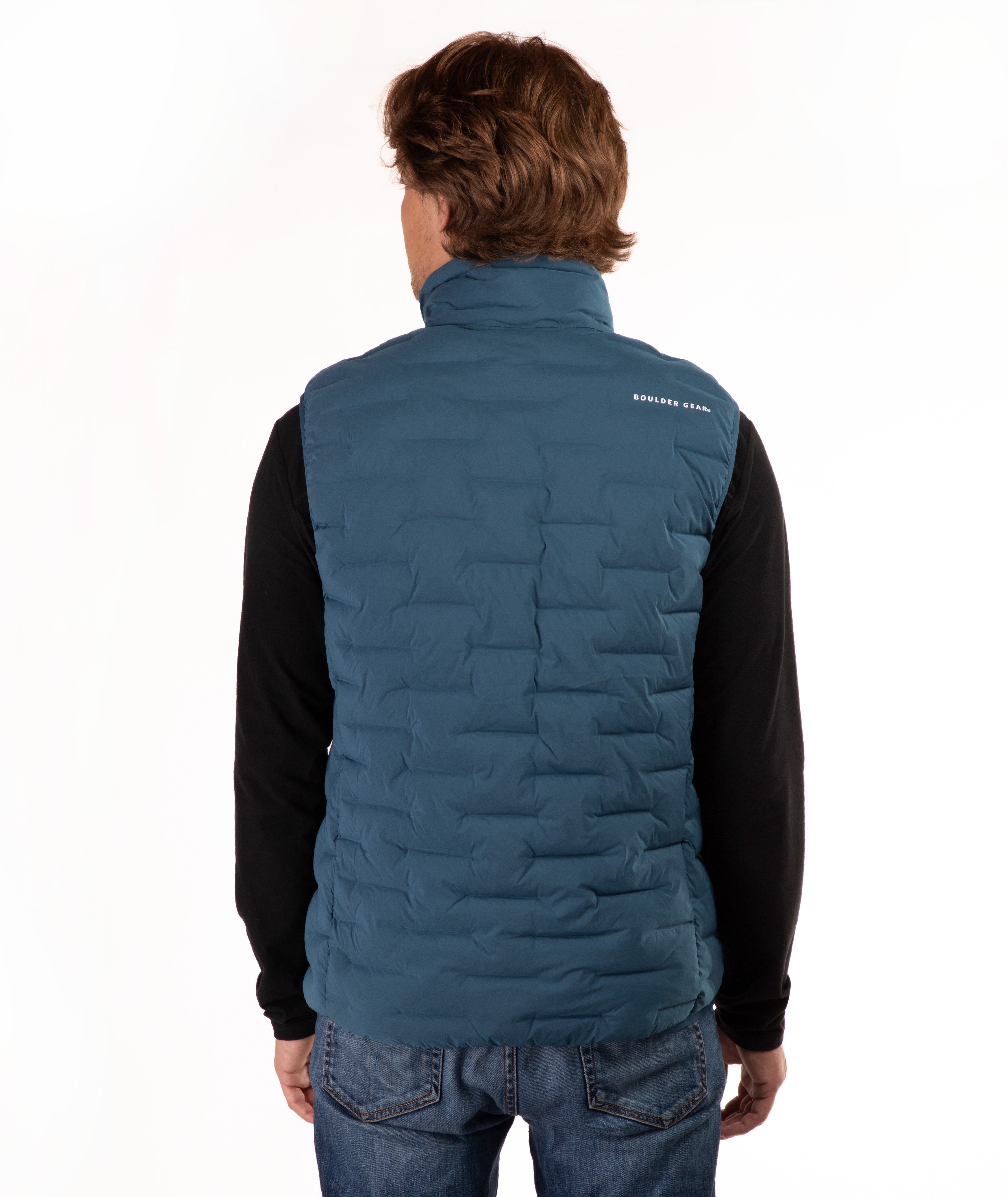 A man viewed from behind wearing a teal blue quilted puffer vest over a black long-sleeve shirt and blue jeans, against a plain white studio background.