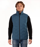 A man wearing a teal blue quilted puffer vest over a black long-sleeve shirt and blue jeans, standing against a plain white studio background.