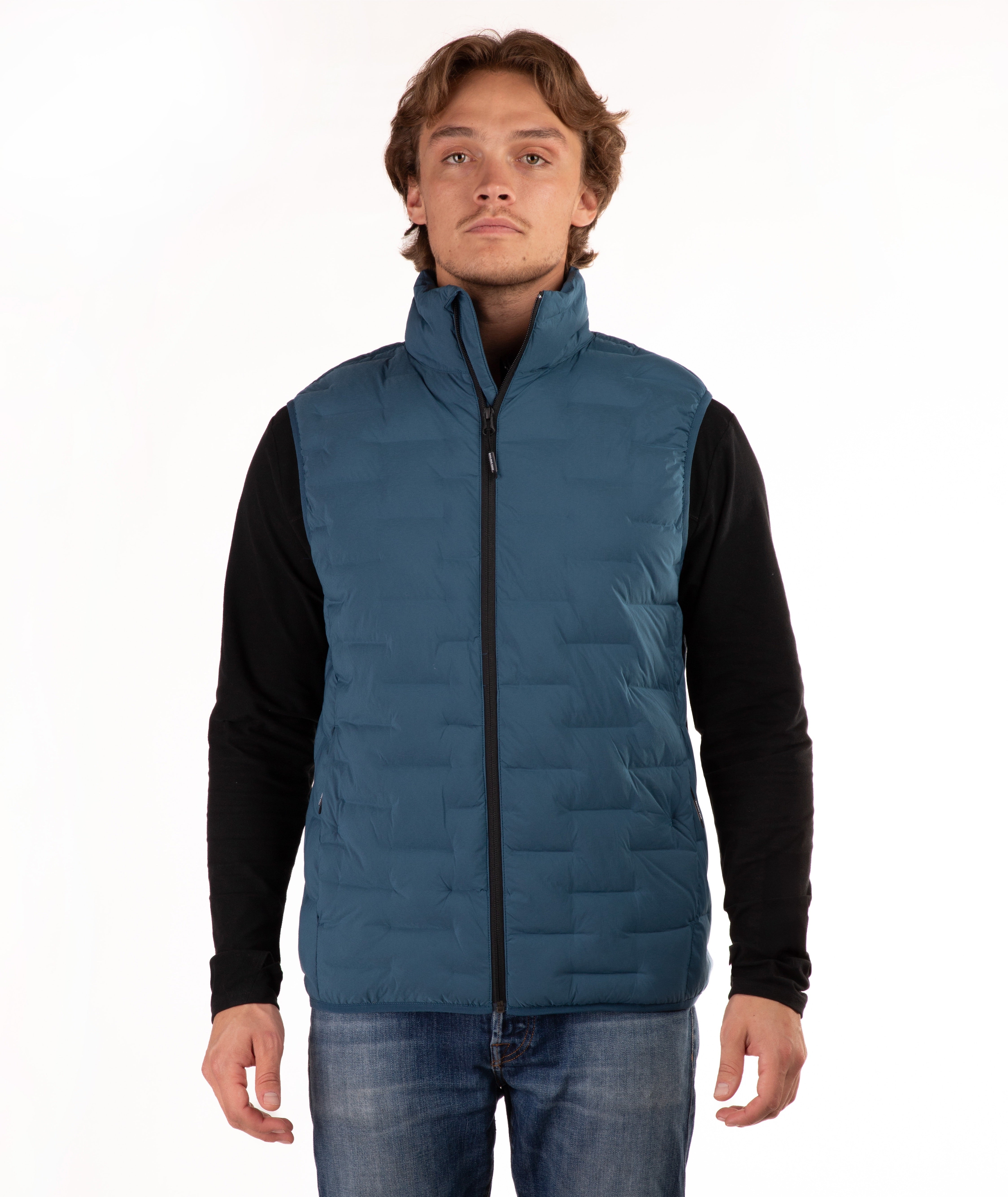 A man wearing a teal blue quilted puffer vest over a black long-sleeve shirt and blue jeans, standing against a plain white studio background.
