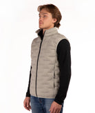 A man wearing a light beige quilted puffer vest over a black long-sleeve shirt and blue jeans, standing against a plain white background.