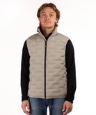A man wearing a light beige quilted puffer vest over a black long-sleeve shirt and blue jeans, standing against a plain white background.