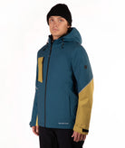 A man wearing a teal and mustard yellow color-blocked ski jacket, a black beanie, and black pants, standing against a plain white studio background.