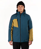 A man wearing a teal and mustard yellow color-blocked ski jacket, a black beanie, and black pants, standing against a plain white studio background.