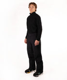 A man wearing a black long-sleeve half-zip athletic top and black ski pants, standing against a solid white studio background.