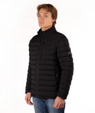 A man wearing a black puffer jacket with a stand-up collar and zippered front, paired with blue jeans, photographed against a plain white background.