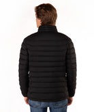 A man viewed from the back wearing a black horizontally quilted puffer jacket and blue jeans, standing against a plain white studio background.