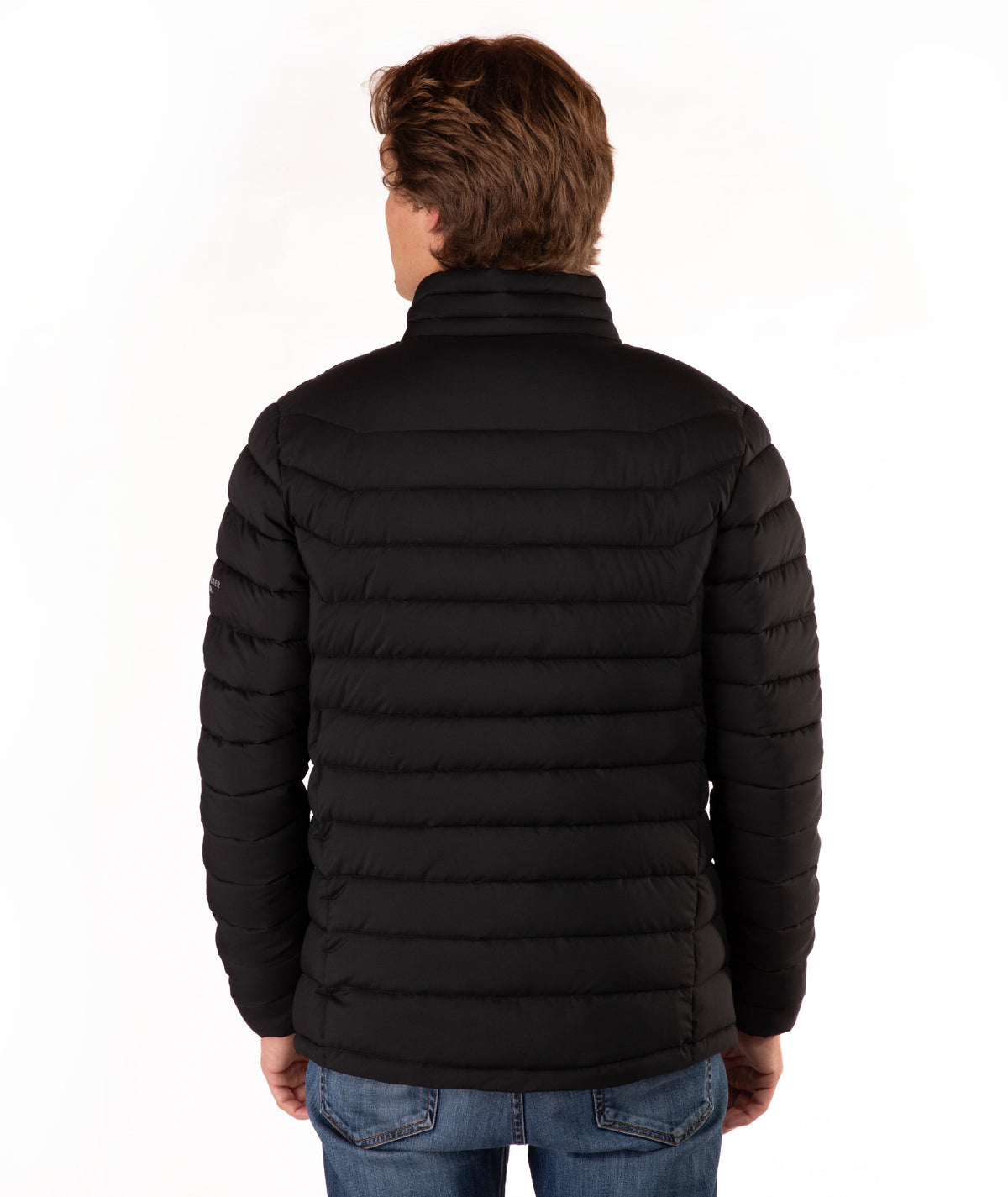A man viewed from the back wearing a black horizontally quilted puffer jacket and blue jeans, standing against a plain white studio background.