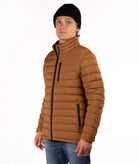 A man wearing a brown puffer jacket with a high collar, black zipper details, a black beanie, and blue jeans, photographed against a plain white background.