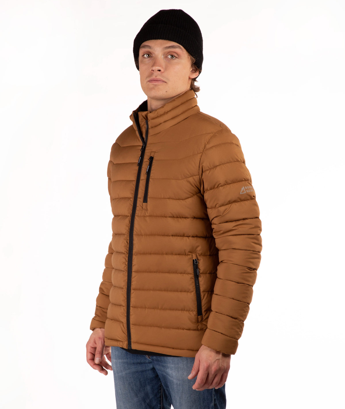 A man wearing a brown puffer jacket with a high collar, black zipper details, a black beanie, and blue jeans, photographed against a plain white background.