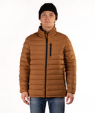 A man wearing a brown puffer jacket with a high collar, black zipper details, a black beanie, and blue jeans, photographed against a plain white background.