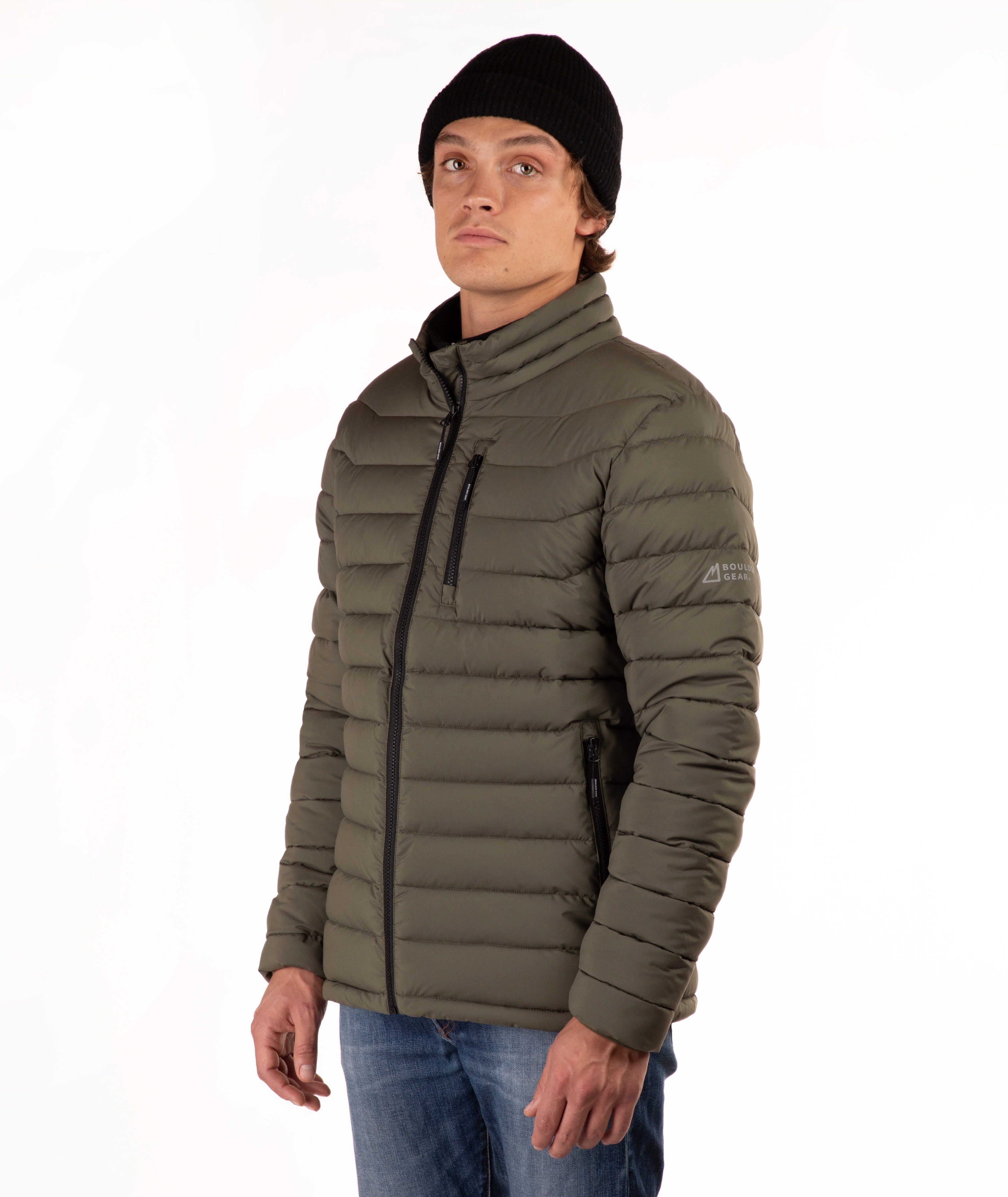 A man wearing a dark olive green puffer jacket with a black zipper, a black beanie, and blue jeans, standing against a plain white background.