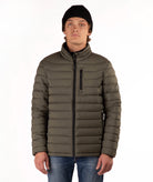 A man wearing a dark olive green puffer jacket with a black zipper, a black beanie, and blue jeans, standing against a plain white background.