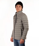 A man wearing a taupe quilted puffer jacket with a high collar, black zipper details, a black beanie, and blue jeans, standing against a plain white background.