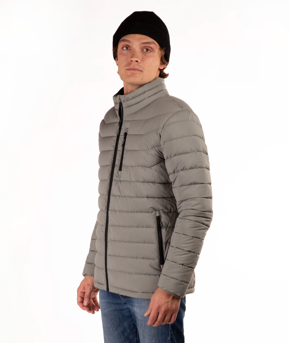 A man wearing a taupe quilted puffer jacket with a high collar, black zipper details, a black beanie, and blue jeans, standing against a plain white background.