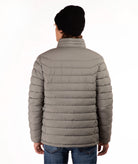 A person shown from the back wearing a taupe quilted puffer jacket, a black knit beanie, and blue jeans against a plain white studio background.